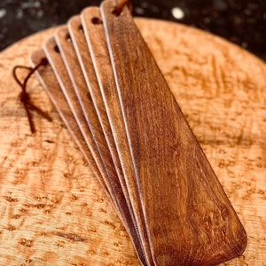 May include: A set of five wooden serving spatulas with a brown leather strap. The spatulas are stacked on top of each other and have a smooth, polished finish.