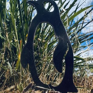 Puede incluir: Una decoración de jardín de metal negro en forma de silueta de pingüino. El pingüino está de pie en un lecho de hierba verde alta.
