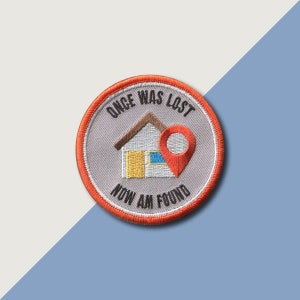 Op de afbeelding: Geborduurde patch met een huis en een rode locatiepin. De tekst "ONCE WAS LOST NOW AM FOUND" staat in een cirkelvormig ontwerp. De patch heeft een oranje rand en een grijze achtergrond.