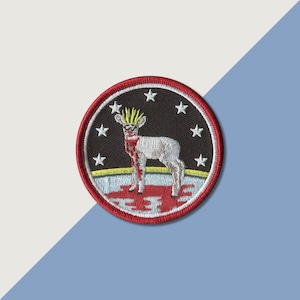 May include: A red, white, and blue embroidered patch with a white lamb with a crown of thorns and blood dripping from its neck. The lamb is standing on a red, white, and blue field with stars.