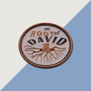 Può includere: Una toppa ricamata bianca con cuciture marroni. La toppa presenta un albero con radici e il testo "THE ROOT of DAVID".