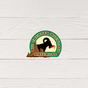 May include: A green circle logo with the text "Big Thunder Mountain Railroad" in brown lettering. Inside the circle is a brown silhouette of a mountain with a black mountain goat holding a red dynamite stick.