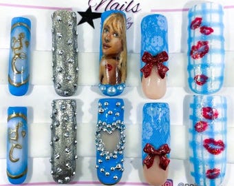 Puede incluir: Diez uñas postizas con diversos diseños. Las uñas presentan tonos azules, plateados y nude, adornadas con pedrería, lazos y un retrato. Algunas uñas tienen un patrón de cuadros o un diseño de encaje. La imagen incluye el texto "Nails" y un perfil de redes sociales.