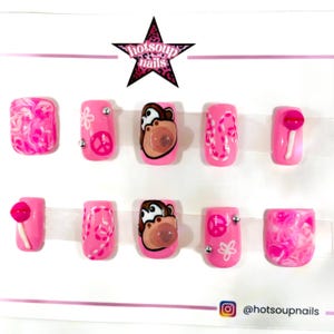 May include: A set of ten pink press-on nails with various designs. Some nails feature a monkey face, peace signs, and swirls. Others have lollipop accents. The brand name "hotsoupnails" is visible on a star-shaped logo.