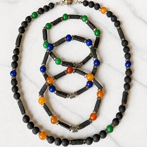 Könnte beinhalten: Eine Halskette und zwei Armbänder mit schwarzen Perlen und zylindrischen Akzenten. Der Schmuck enthält farbige Perlen in Grün, Blau, Orange und Gelb. Die Halskette hat einen Magnetverschluss.
