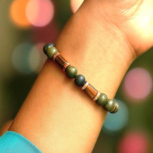 May include: A beaded bracelet featuring olive green and dark gray beads, interspersed with copper-colored cylindrical beads and silver-toned spacers. The bracelet is displayed on a wrist, with a blurred background of colorful bokeh lights.