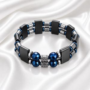 May include: A black and silver bracelet with blue accents. The bracelet features rectangular and cylindrical metal pieces, interspersed with faceted blue beads. The design includes a decorative silver clasp, set against a white satin background.