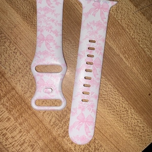 Op de afbeelding: Een paar roze en witte gepatenteerde horlogebandjes met een bloemenmotief en roze strikken. De bandjes zijn gemaakt van siliconen en zijn compatibel met Apple Watches.