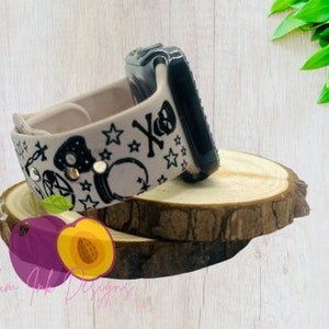 May include: A light pink silicone watch band with a black and white design featuring stars, a skull, and other symbols. The band is designed for a smartwatch and has a black metal clasp.