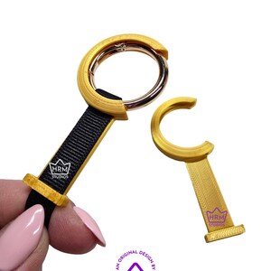 May include: Two gold-colored keychains with black straps and a silver clasp. One keychain is attached to a black strap, and the other is a separate piece. The keychains have the text "HRM STUDIOS" on them. The image also includes the text "AN ORIGINAL DESIGN BY HRM STUDIOS."