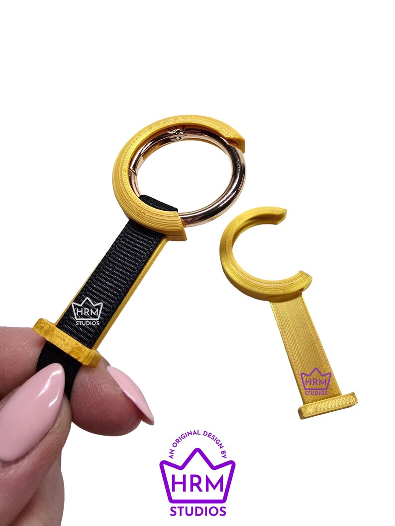 May include: Two gold-colored keychains with black straps and a silver clasp. One keychain is attached to a black strap, and the other is a separate piece. The keychains have the text "HRM STUDIOS" on them. The image also includes the text "AN ORIGINAL DESIGN BY HRM STUDIOS."