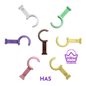 Puede incluir: Una colecci&oacute;n de ganchos en forma de C de colores, en blanco, verde, morado, marr&oacute;n, amarillo y rosa. Cada gancho tiene una base rectangular con un logotipo de corona y el texto "HAS". La imagen tambi&eacute;n incluye el texto "AN ORIGINAL DESIGN BY HRM STUDIOS".