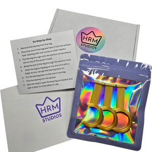 Puede incluir: Un paquete de producto con tres llaveros dorados en una bolsa hologr&aacute;fica. El paquete incluye una caja blanca y una tarjeta con instrucciones y el texto "Go Step-by-Step". El logotipo de HRM Studios es visible en la caja y la tarjeta.