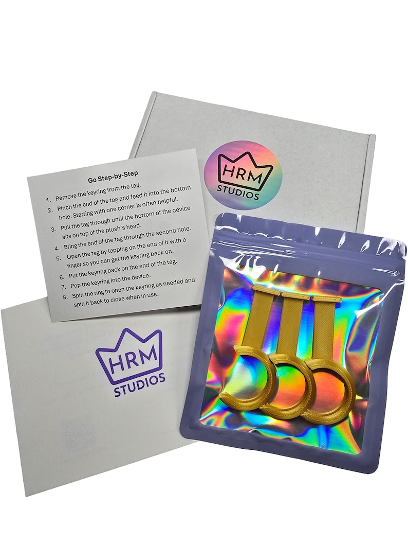 Puede incluir: Un paquete de producto con tres llaveros dorados en una bolsa hologr&aacute;fica. El paquete incluye una caja blanca y una tarjeta con instrucciones y el texto "Go Step-by-Step". El logotipo de HRM Studios es visible en la caja y la tarjeta.