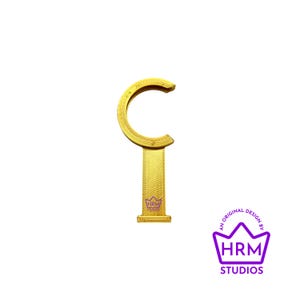 Puede incluir: Un gancho de cortina dorado con una barra vertical texturizada y una parte superior curvada. El gancho tiene una peque&ntilde;a base rectangular y un dise&ntilde;o decorativo. El logotipo "HRM STUDIOS" est&aacute; en la esquina inferior derecha.