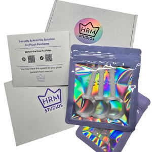 May include: A product package from HRM Studios, featuring a white box and a card with the text "Security & Anti-Flip Solution for Plush Pendants." The package includes three silver key-shaped items in a holographic bag. The HRM Studios logo is visible.