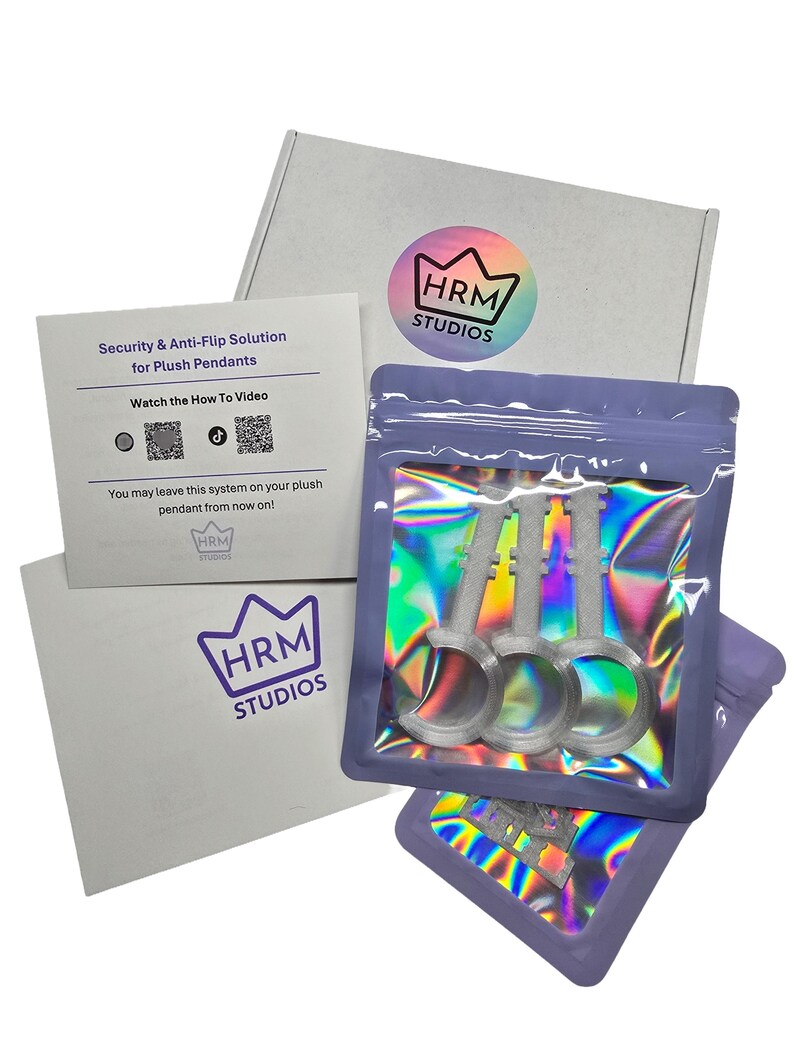 May include: A product package from HRM Studios, featuring a white box and a card with the text "Security & Anti-Flip Solution for Plush Pendants." The package includes three silver key-shaped items in a holographic bag. The HRM Studios logo is visible.