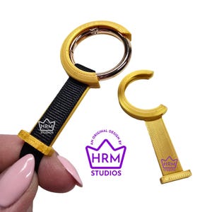 Puede incluir: Dos llaveros dorados con correas negras. Un llavero tiene un cierre circular y el otro un diseño en forma de C. Los llaveros tienen el texto "HRM STUDIOS" impreso. Los llaveros están sobre un fondo blanco.