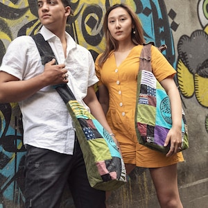 May include: Two people are standing in front of a graffiti wall. The person on the left is wearing a white shirt and black pants and carrying a multi-colored patchwork bag. The person on the right is wearing a yellow dress and carrying a similar multi-colored patchwork bag.