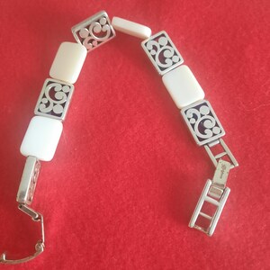 May include: Silver-toned bracelet with rectangular ivory-colored links and decorative openwork squares. The bracelet is displayed against a red background, showcasing its intricate design and clasp.