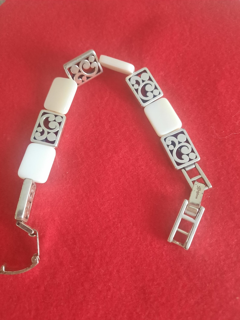 May include: Silver-toned bracelet with rectangular ivory-colored links and decorative openwork squares. The bracelet is displayed against a red background, showcasing its intricate design and clasp.