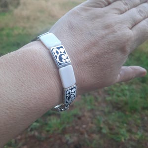 May include: A silver bracelet featuring rectangular white segments and decorative silver and black squares. The bracelet is shown on a wrist, with the clasp visible. The design includes a repeating pattern of black swirls within the silver squares.
