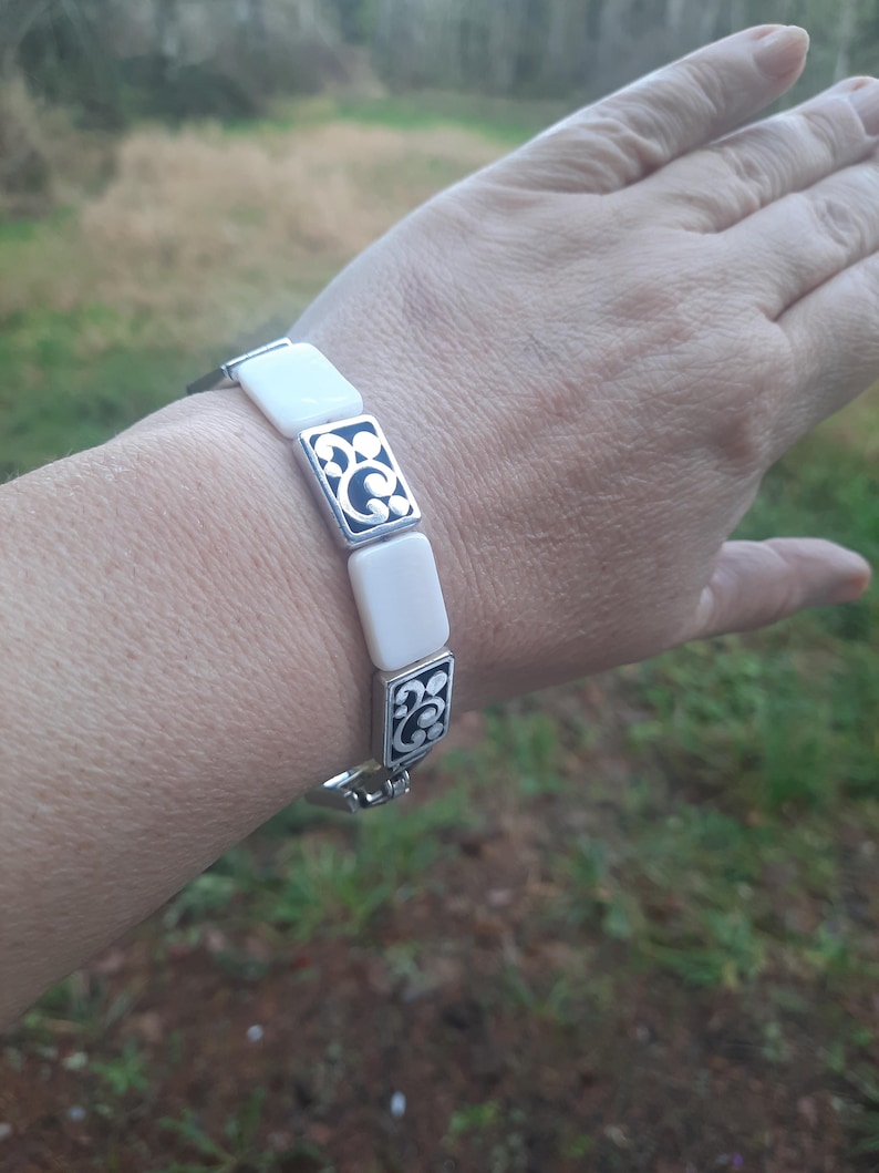 May include: A silver bracelet featuring rectangular white segments and decorative silver and black squares. The bracelet is shown on a wrist, with the clasp visible. The design includes a repeating pattern of black swirls within the silver squares.
