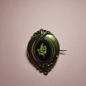 May include: A gold oval brooch with a black cameo featuring a green floral design with red accents.