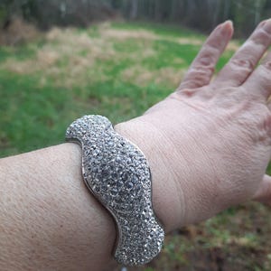May include: Silver-toned cuff bracelet with a bird-like design, fully encrusted with clear, sparkling crystals. The bracelet is worn on a wrist, showcasing its curved shape and reflective surface. The background is a blurred outdoor scene.