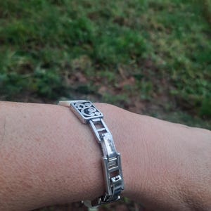 May include: Silver-toned bracelet with a decorative square centerpiece and rectangular links. The centerpiece features a black design. The bracelet is worn on a wrist, with a blurred green background.