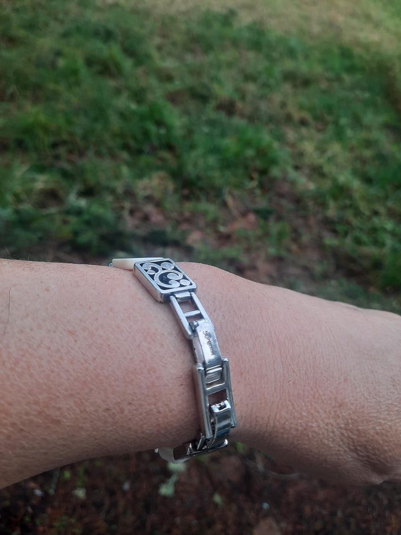 May include: Silver-toned bracelet with a decorative square centerpiece and rectangular links. The centerpiece features a black design. The bracelet is worn on a wrist, with a blurred green background.