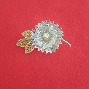 May include: A silver-toned flower brooch with a central iridescent rhinestone. The brooch features a layered design with heart-shaped petals and a stem with two gold-colored leaves. The background is a solid red.