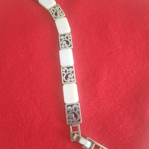May include: A silver-toned bracelet with rectangular white segments and decorative filigree squares. The bracelet is displayed on a red surface. The clasp is open, showing the bracelet's construction.