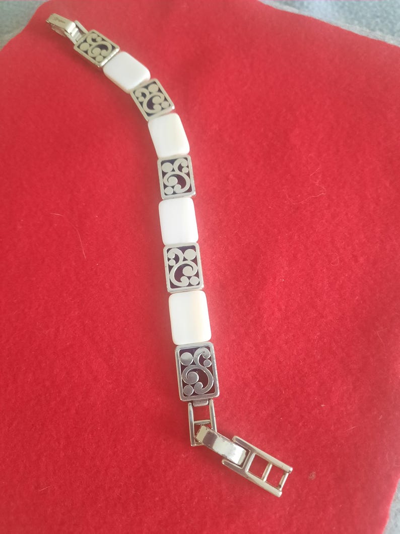 May include: A silver-toned bracelet with rectangular white segments and decorative filigree squares. The bracelet is displayed on a red surface. The clasp is open, showing the bracelet's construction.