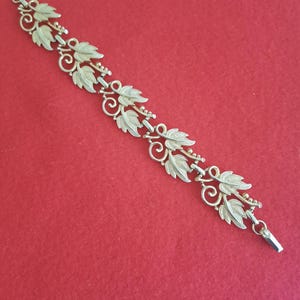 Puede incluir: Un brazalete dorado con un diseño de hojas y espirales. El brazalete presenta una serie de hojas estilizadas conectadas por delicados eslabones y pequeños detalles de cuentas. El cierre es de diseño simple y rectangular.