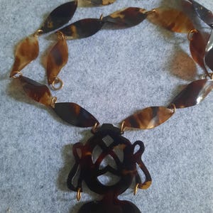 May include: A necklace featuring a pendant with a stylized figure and leaf-shaped links. The necklace is made of brown and tan materials, with gold-colored connectors. The pendant has a geometric design.