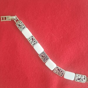 May include: Silver-toned bracelet with rectangular white segments and decorative filigree accents. The bracelet is laid out diagonally against a red background, showcasing its design and clasp.