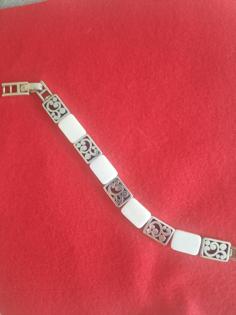 May include: Silver-toned bracelet with rectangular white segments and decorative filigree accents. The bracelet is laid out diagonally against a red background, showcasing its design and clasp.