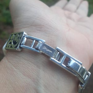 May include: Silver-toned bracelet with a decorative square element featuring an openwork design. The bracelet has a clasp with the brand name "Brighton" engraved. The band appears to be white.
