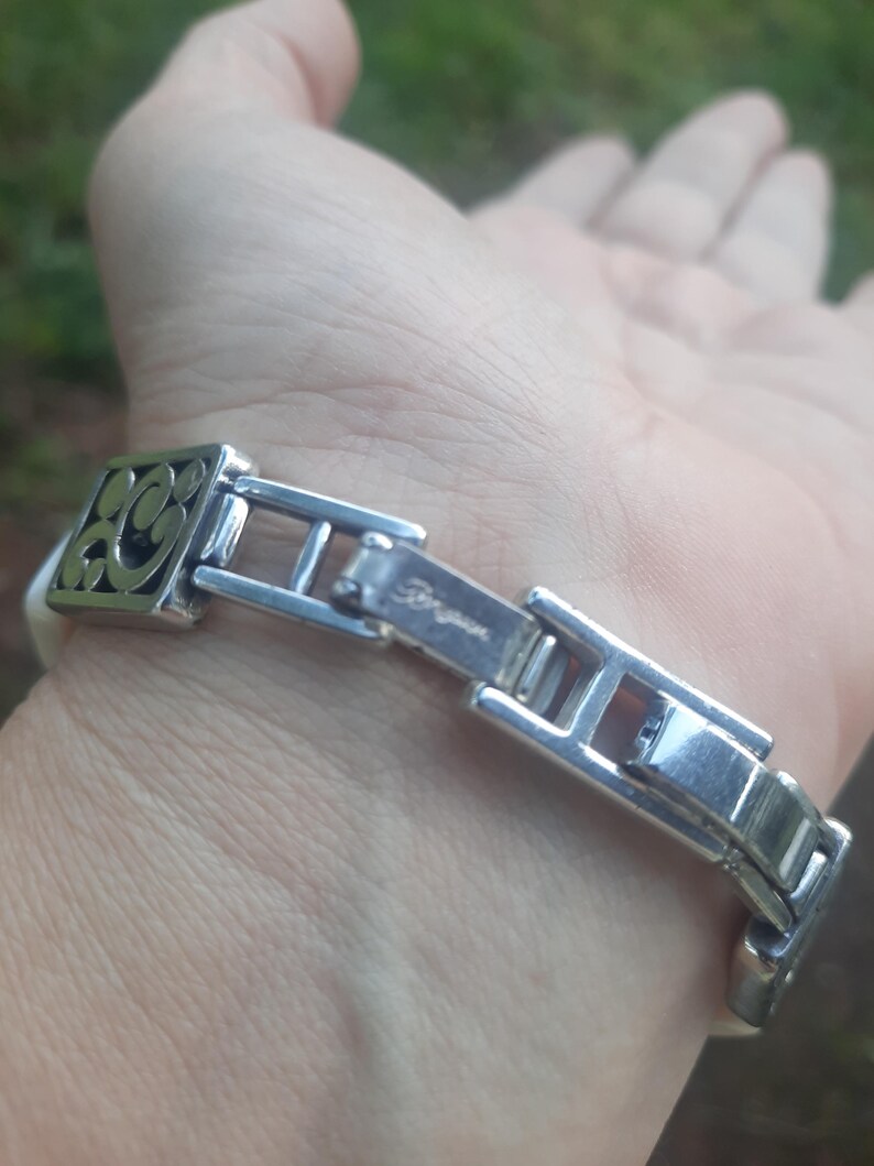 May include: Silver-toned bracelet with a decorative square element featuring an openwork design. The bracelet has a clasp with the brand name "Brighton" engraved. The band appears to be white.