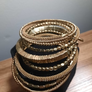 May include: A stack of gold-toned bangles with different textures and patterns. The bangles are arranged in a circular shape and are stacked on top of each other.
