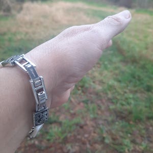 May include: A silver-toned bracelet with a decorative design. The bracelet features rectangular and square links with an ornate, floral-like pattern. The bracelet is on a wrist, with a blurred outdoor background.