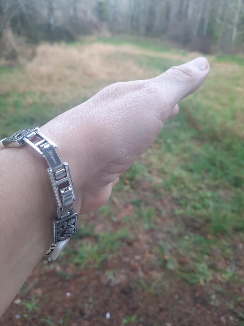 May include: A silver-toned bracelet with a decorative design. The bracelet features rectangular and square links with an ornate, floral-like pattern. The bracelet is on a wrist, with a blurred outdoor background.