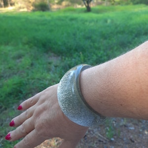 May include: A clear, wide bangle bracelet with silver glitter embedded throughout. The bracelet is worn on a wrist with red nail polish. The background is a natural outdoor setting with green grass and trees.