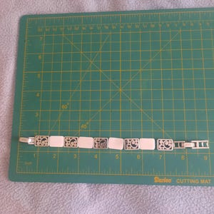 May include: A silver-toned bracelet with rectangular white segments and decorative filigree links. The bracelet is laid on a green cutting mat with inch measurements.