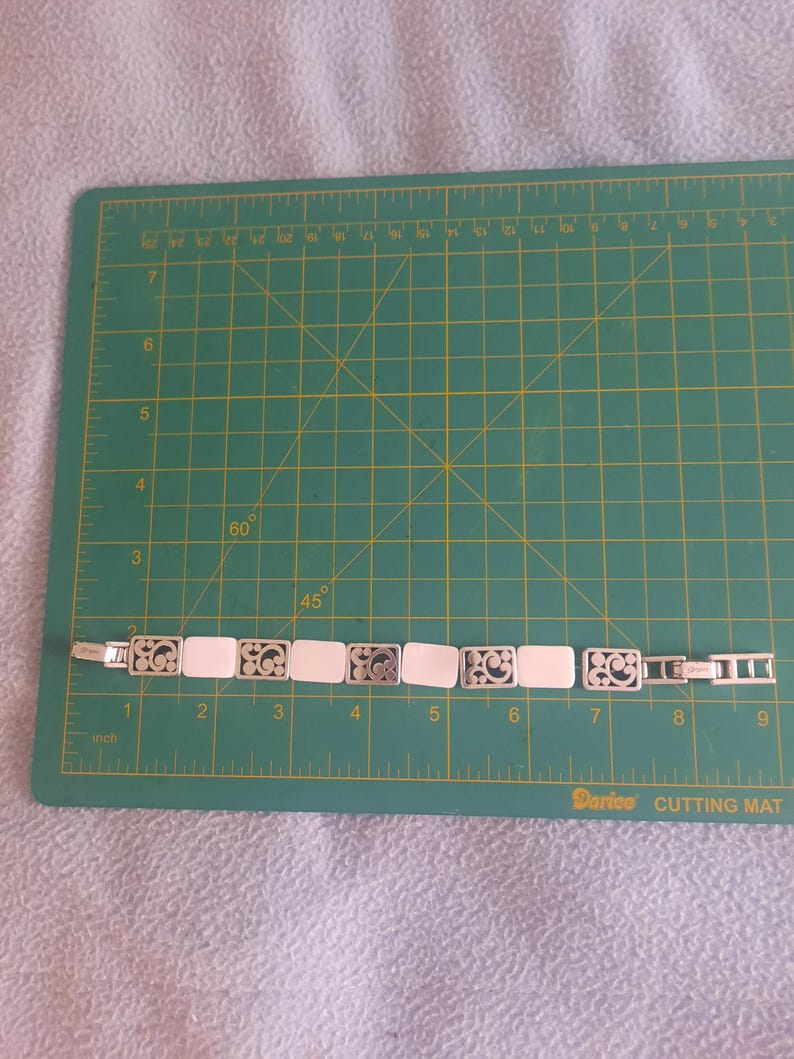 May include: A silver-toned bracelet with rectangular white segments and decorative filigree links. The bracelet is laid on a green cutting mat with inch measurements.