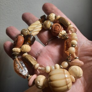 May include: A multi-strand necklace with brown, beige, and gold beads. The necklace features a variety of textures, including smooth, carved, and textured beads. The necklace is made of wood, glass, and metal.