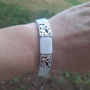 May include: A white and silver bracelet with a rectangular design. The bracelet features alternating white rectangular links and silver links with a black, swirling pattern. The bracelet is worn on a wrist.