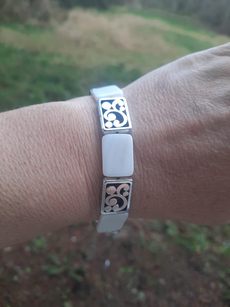 May include: A white and silver bracelet with a rectangular design. The bracelet features alternating white rectangular links and silver links with a black, swirling pattern. The bracelet is worn on a wrist.
