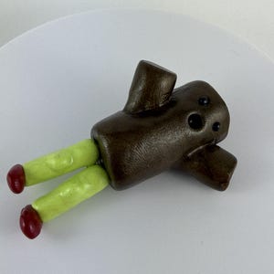 May include: A quirky, handmade figurine. It has a brown, cylindrical body with two small arms and three black dots. The figurine has two green legs with red tips. It is lying on a white surface.
